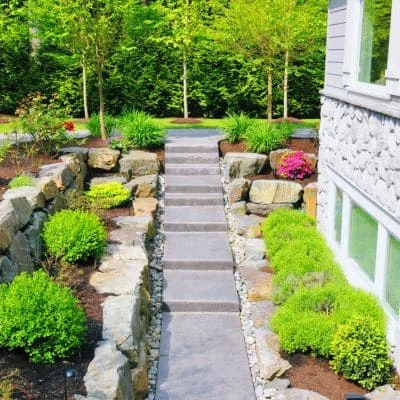 Stone retaining walls, concrete pathway, and tiered plantings with green shrubs and pink flowers