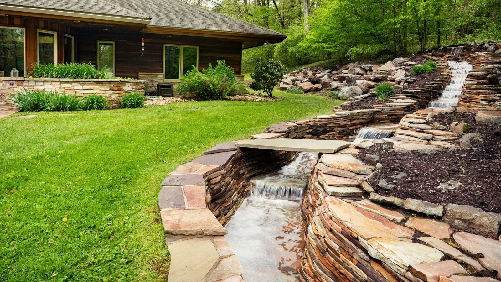 Stacked stone creek bed with cascading water, flagstone stepping stones, and layered retaining walls