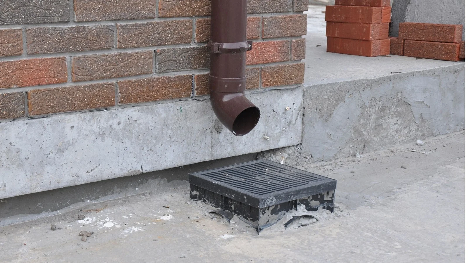 Brown downspout with catch basin and grate on concrete foundation near brick wall
